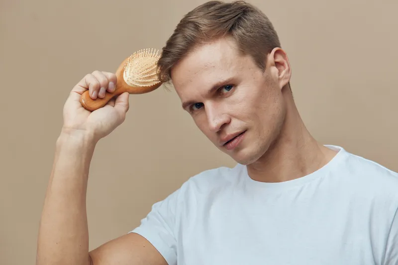 Young caucasian man brushing hair with wooden brush white t-shirt closeup neutral beige background hair care personal hygiene concept male portrait