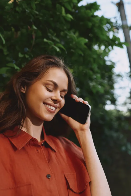 Woman in outdoor lifestyle scene smiles while talking on a smartphone, wearing a rust shirt, standing among green trees, relaxed casual portrait conveying everyday communication