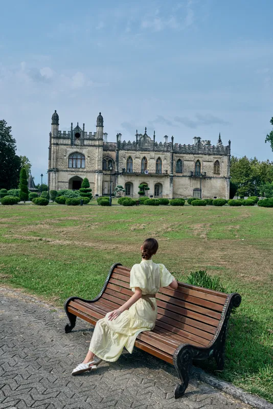 A woman in a pale dress sits on a wooden bench in a sunny park, with a historic castle like building visible in the background, creating a calm scenic travel moment.