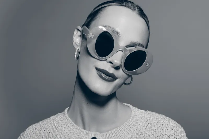 Elegant white woman with pale skin wears large round sunglasses and hoop earrings, posing confidently in studio. Concept for fashion and style.