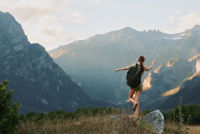 A young woman with a backpack strides through a grassy meadow toward dramatic mountains, radiating adventure, freedom, and focus on the trail ahead.