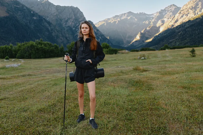 A young female traveler stands in a green mountain meadow, gripping a trekking pole and camera, dressed in a dark jacket and shorts, with dramatic alpine peaks in the backdrop.