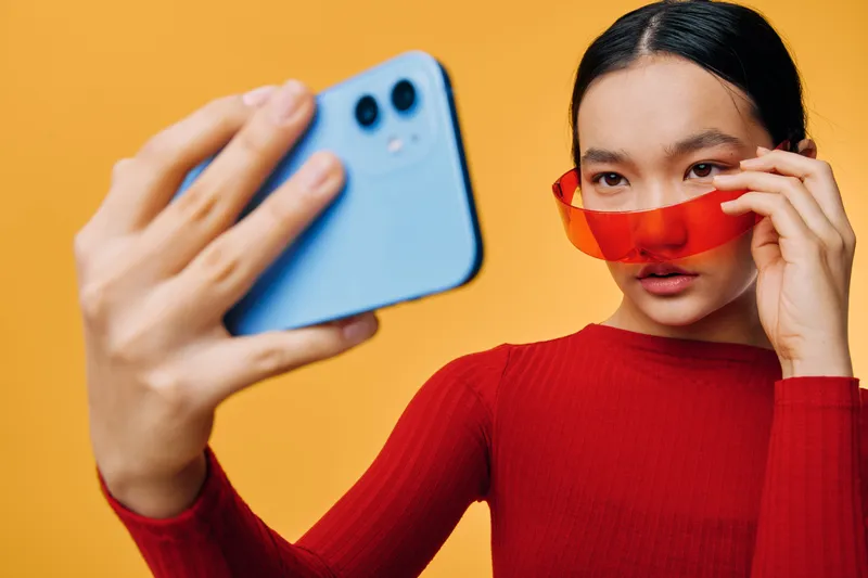 Young woman in red sweater and red sunglasses taking selfie with smartphone on bright yellow background. Female posing with phone for social media photo or digital portrait closeup.
