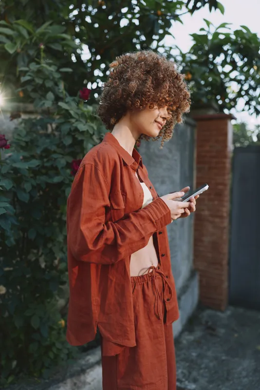 A woman with curly hair wearing rust loungewear checks her phone outdoors, leaning against a wall in a sunny garden. Casual lifestyle moment highlighting comfort and personal style