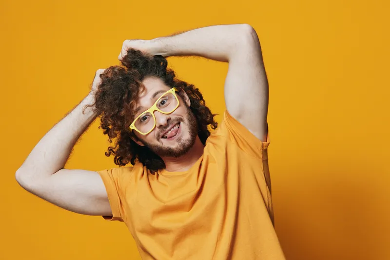 Young man with curly hair and yellow glasses, smiling against a vibrant yellow background in casual summer attire, reflecting joy and positivity