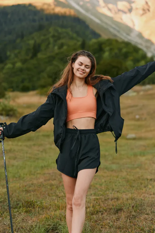 A cheerful young woman stands outdoors in a mountain meadow, wearing an athletic jacket, shorts, and a crop top. She smiles warmly, arms open, enjoying fresh air and a sense of freedom.