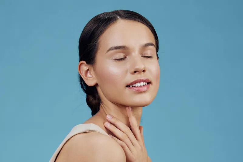 Young woman with closed eyes and gentle smile, wearing neutral top, against pastel blue background, conveying serenity and beauty with natural makeup
