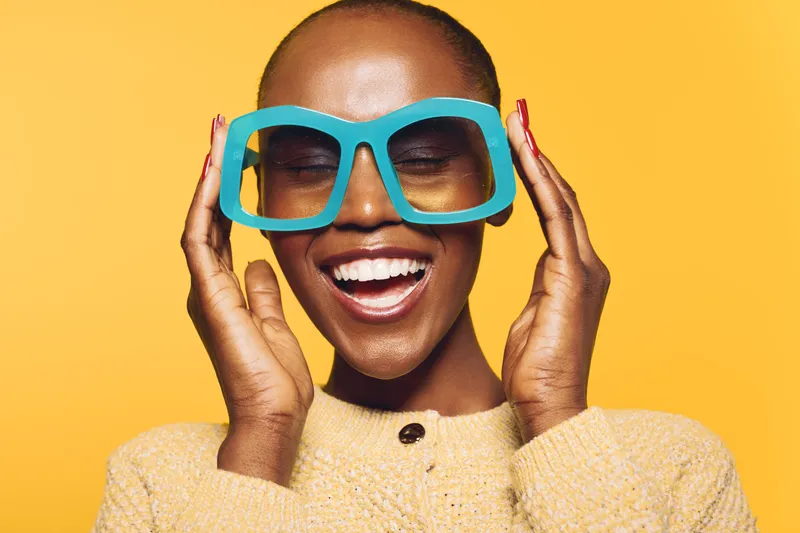 Happy African woman with dark skin wearing large blue glasses and a yellow sweater smiles with closed eyes on a bright yellow background. Concept of joy, fashion, and beauty.