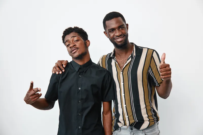 Two African American men with dark skin posing together smiling and showing thumbs up and hand gesture on white background. Friends, casual, happiness and confidence.