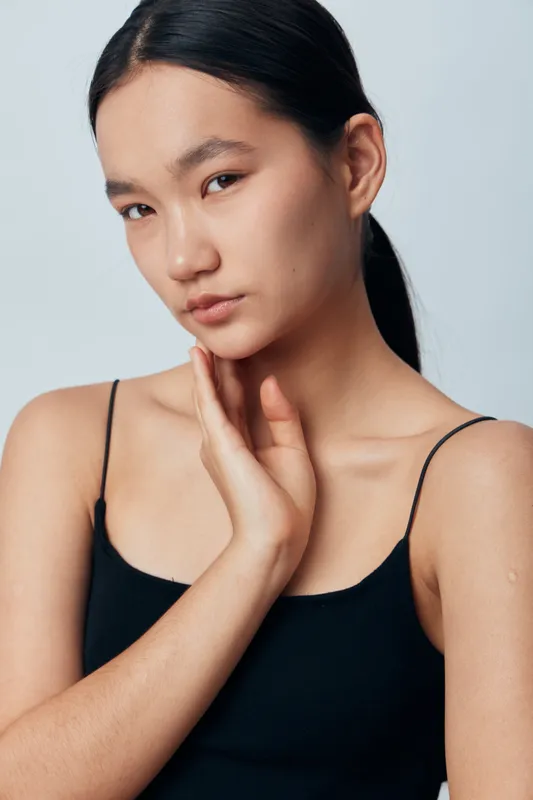 Young woman with black hair and natural makeup posing with hand near neck wearing black tank top on plain background. Studio portrait of female model with calm expression and smooth skin.