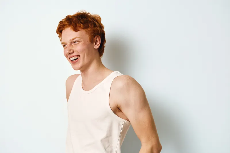 Young man with red hair laughing and wearing white tank top in studio with plain background, showing natural smile and relaxed casual style.