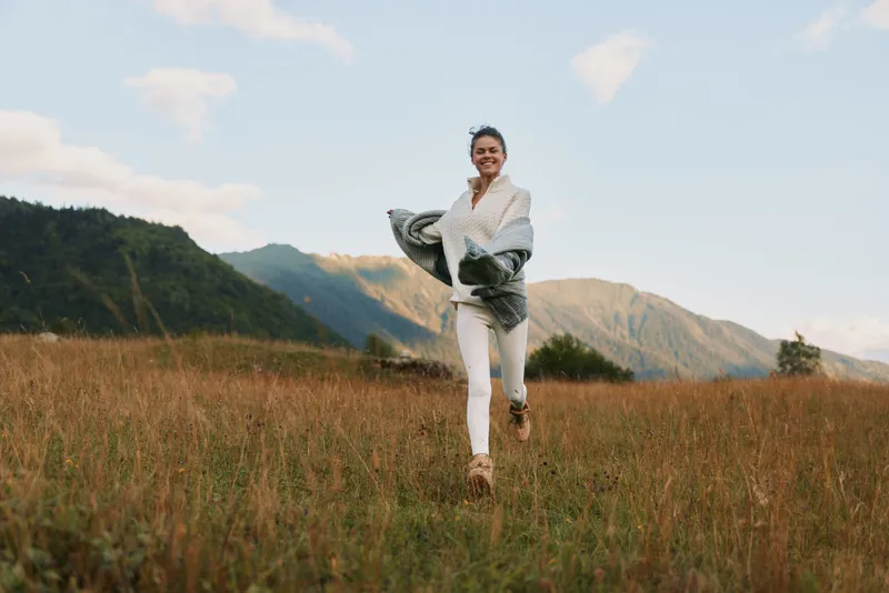 Smiling woman walking through a tall grass field with distant hills and mountains, capturing a relaxed outdoor lifestyle moment in natural light, evoking freedom and wellness.