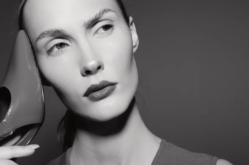 Portrait of a young Caucasian woman with pale skin holding a high heel shoe near her face in a black and white studio shot. Concept of fashion and elegance.