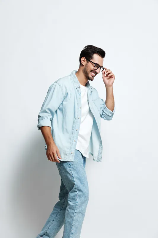 Full body smiling cheerful young man wearing a light blue shirt and jeans, walking while adjusting his glasses, isolated on a plain light background. Fashion lifestyle concept.
