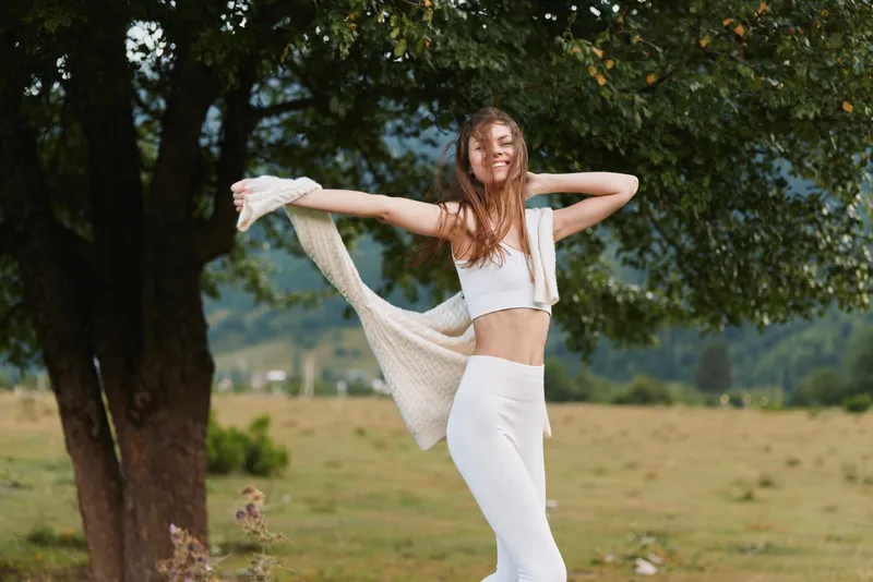 A young woman dances joyfully in a sunny park, wearing a white crop top and white pants with a light scarf, radiating carefree vitality and balance.