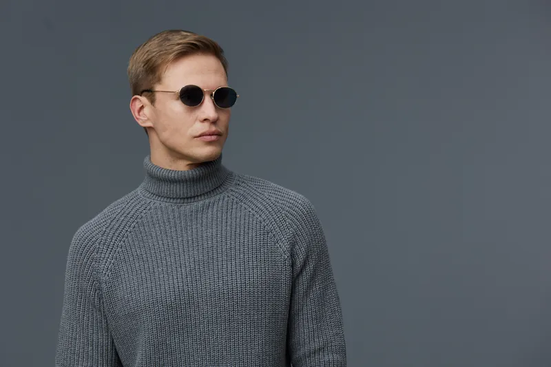 Stylish young man with blonde hair, wearing round sunglasses and a gray turtleneck sweater, posing confidently against a plain gray background. Modern fashion look, casual style, studio portrait. Peop
