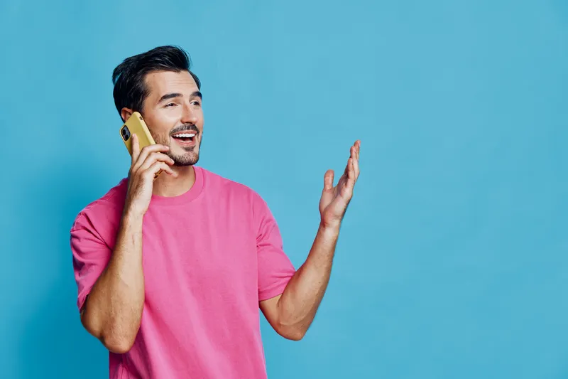 Energetic smiling man wearing pink shirt looks cheerful while talking on a mobile phone against a bright blue background in studio portrait. Communication concept