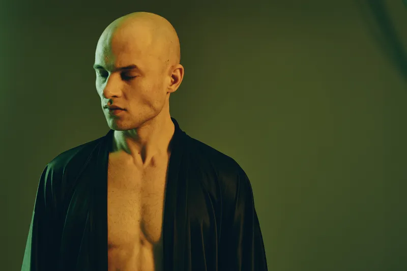 A contemplative man in a dark robe, showcasing a serene expression and bald head. The soft green lighting creates a calm and artistic atmosphere.