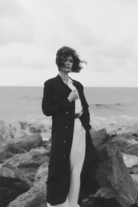 Fashion model stands on a rocky shoreline in a long coat, wind whipping hair, monochrome scene highlighting outerwear, attitude, and coastal mood against the open ocean backdrop