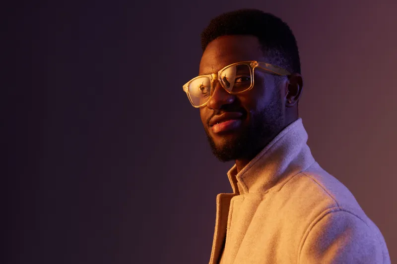 Stylish young African man in a soft beige jacket and yellow glasses against a purple background showcasing confidence and charisma