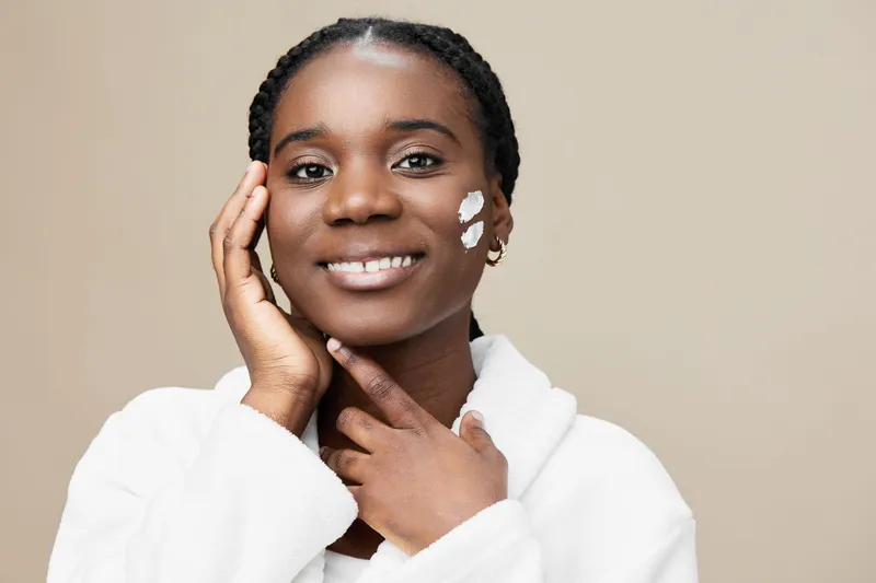 Smiling Black woman applying skincare product on her face, wearing a cozy white robe against a soft beige background, embodying beauty and self care