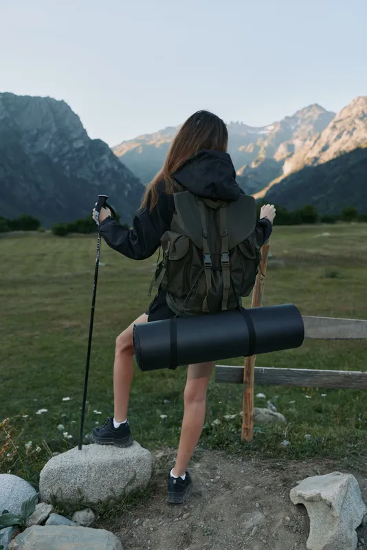 An outdoor traveler stands on a rocky trail, with a large backpack and trekking pole. She faces distant mountains, ready for exploration, and the scene conveys adventure, focus, and freedom.