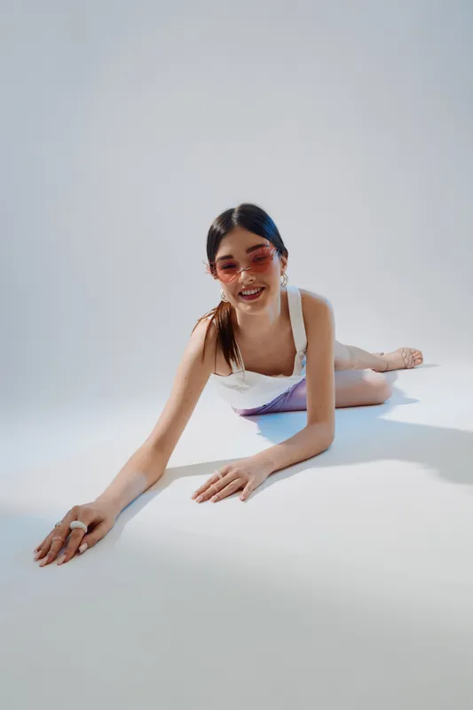 Young woman in white outfit and pink sunglasses lies on floor smiling. Studio shot on white background. Concept of beauty, fashion, and youth lifestyle.