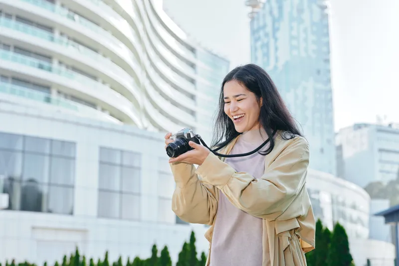 Smiling woman holds camera outdoors in an urban plaza, capturing a candid moment with modern buildings and greenery in the background, showcasing lifestyle photography and travel curiosity