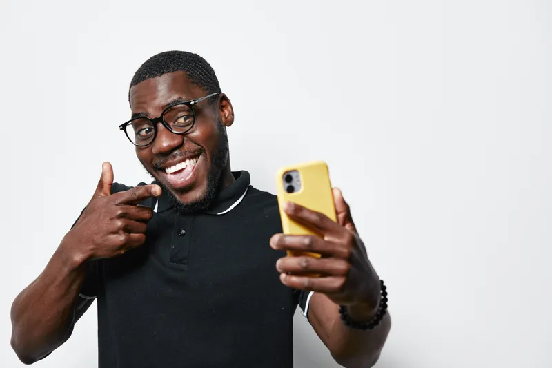 Black man with dark skin and glasses taking selfie with smartphone, smiling and pointing finger, casual style in black polo shirt on white background, happy and confident.