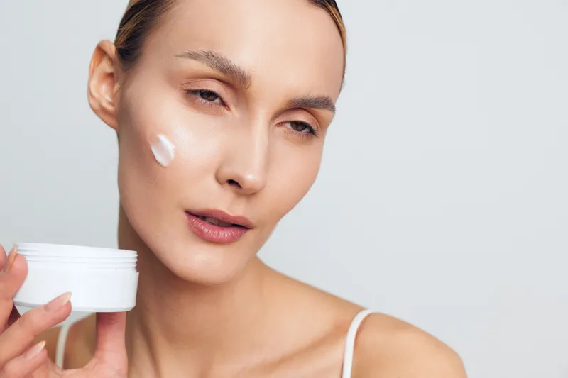 Young woman applies face cream with smooth skin holds white jar skincare product looks relaxed isolated on white background beauty care concept natural makeup routine.