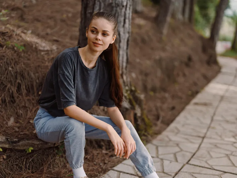 Стильная молодая женщина с длинными волосами, одетая в удобную футболку и джинсы, улыбается, сидя у дорожки в спокойной природной обстановке