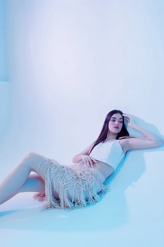 Young woman in a white crop top and fringed skirt poses against a light background. Her long hair flows as she looks thoughtfully to the side. Concept of beauty, fashion, and style.