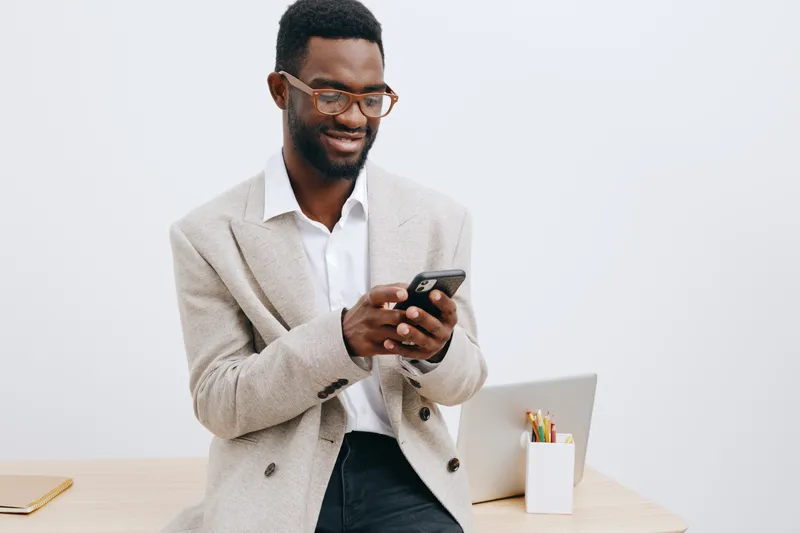 Smiling man using smartphone in office, wearing blazer and white shirt, modern workspace with laptop and stationery, professional and confident demeanor, indoor environment. Business lifestyle concept