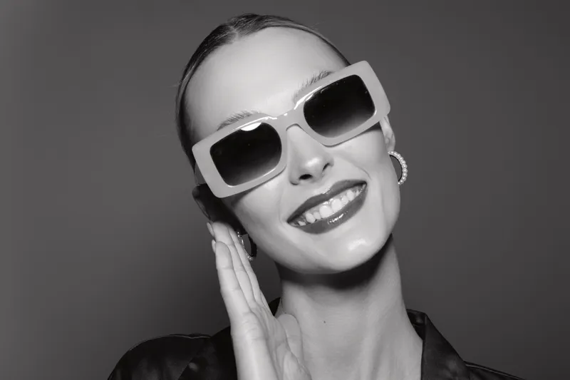 Happy white woman with fair skin and slicked hair wears large sunglasses and smiles confidently on grey background. Concept of fashion, style, and beauty portrait.