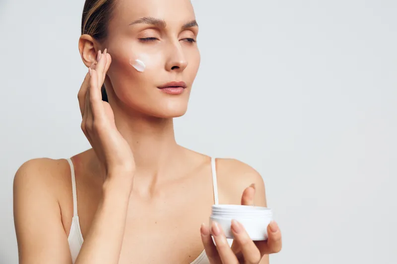 Woman with closed eyes applying white face cream on cheek holding jar. Concept of skincare, beauty routine, self-care, and moisturizing. Soft natural light on plain background.