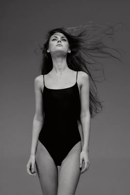 Black and white image of a confident woman with flowing hair wearing a black swimsuit, standing against a plain background.