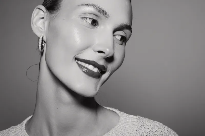 Close-up of a smiling white woman with light skin wearing earrings and white sweater on grey background.