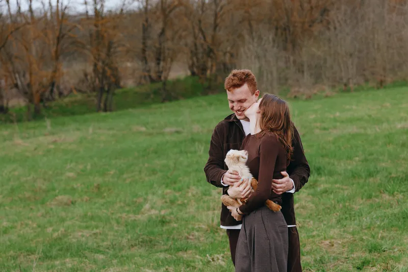 Couple embracing a small goat kid in a grassy field, autumn trees in the background, warm candid moment of affection and rural lifestyle, soft natural light highlights their smiles.