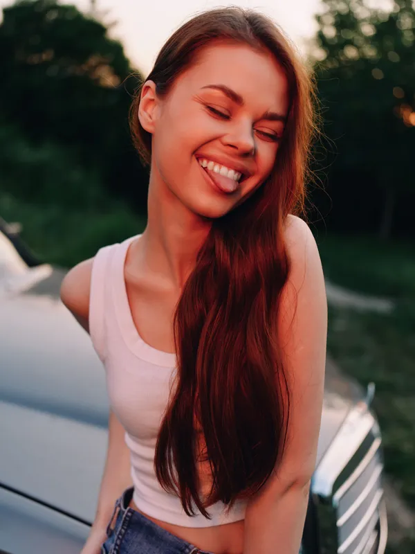 Young woman with a joyful expression in a casual outfit poses outdoors near a car during sunset, highlighting a carefree summer vibe.