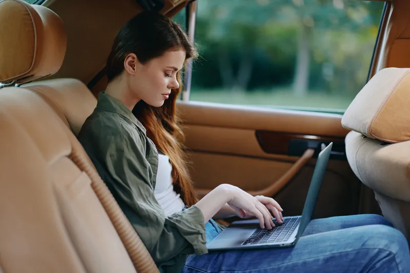 Young woman working on a laptop in the back seat of a car, showcasing a modern lifestyle with a focus on productivity and comfort.