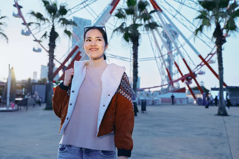 Woman wearing a brown and cream jacket at an outdoor amusement park with a ferris wheel in the background, showcasing fashion, leisure, and urban exploration under sunny sky