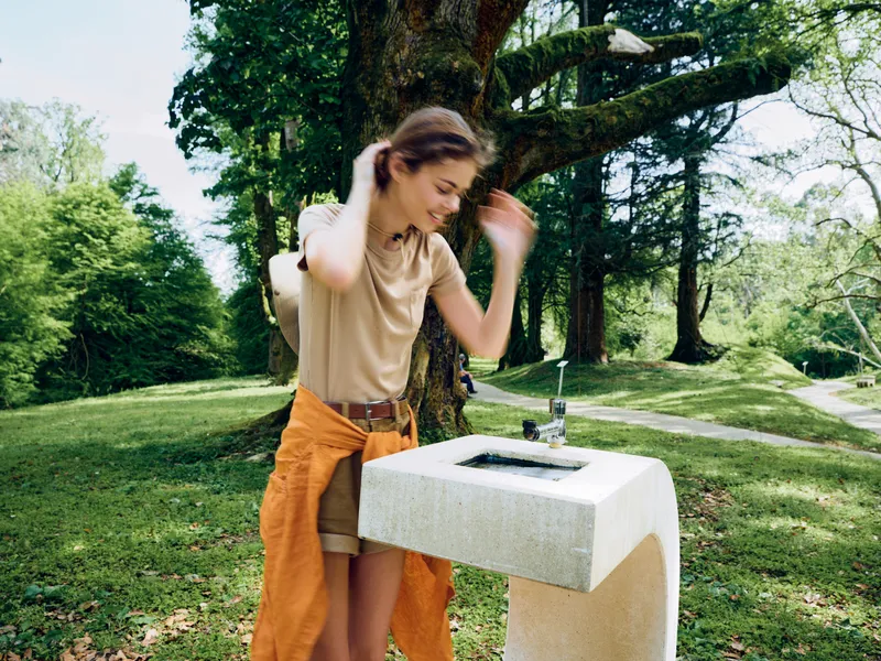 Woman at a fountain in a park with a backpack, enjoying nature in summer on a sunny day. Young casual traveler adjusts hair by the drinking fountain with a relaxed smile.