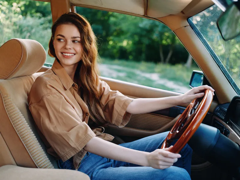 Young woman driving a vintage car, smiling joyfully, wearing a casual beige shirt and blue jeans, surrounded by a lush green forest background.