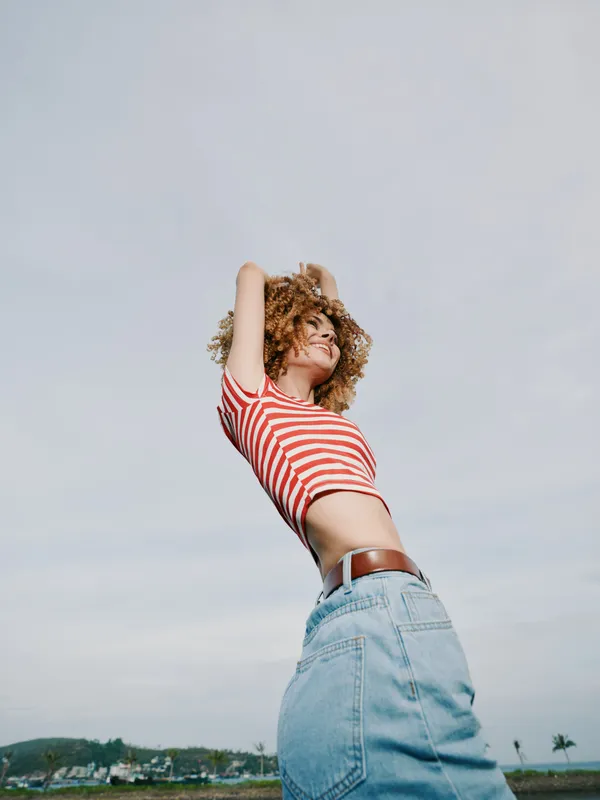 Young woman outdoors wearing a red white striped top and high waisted jeans, leaning back with arms raised, capturing a carefree moment against a bright sky and distant landscape.