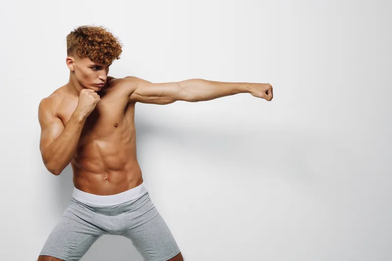 Athletic muscular young man shirtless training boxing at gym, throwing punch with focused expression against neutral background for fitness and combat workout.