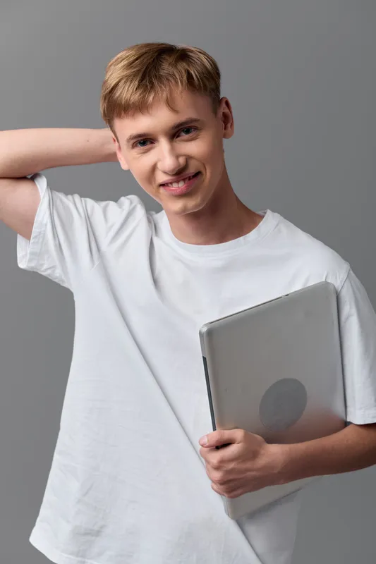 Teenage boy in a white t shirt holding a laptop, smiling and relaxed, teenage student ready for education and learning, ideal for online schooling and technology themed stock in a casual studio