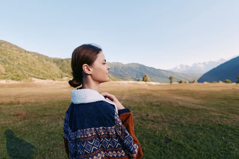 Woman in patterned jacket standing in meadow, portrait and profile view with mountains and landscape in background. Travel scene with serene nature, grass field and clear sky.