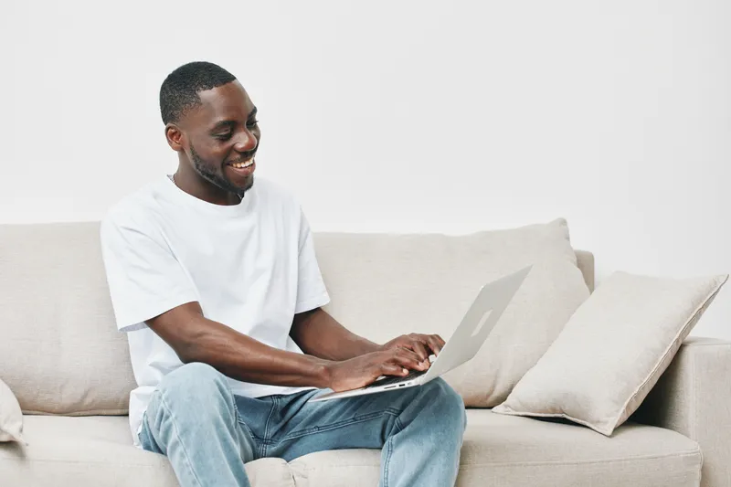 Happy Black man working on laptop in casual outfit at home, smiling on couch, showcasing comfort and productivity in a minimalist setting