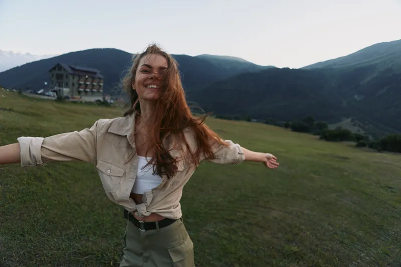 Cheerful woman outdoors, embracing a sunlit moment in a green field, arms open, carefree smile, natural breeze, mountain backdrop, vibrant natural colors with warmth.
