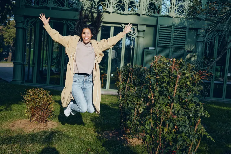Child girl jumping outdoors in garden with coat, expressing happiness and joy, sunlit yard scene, playful moment captured for lifestyle, outdoor activity and happiness